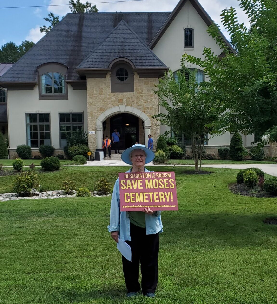 A white woman in a blue hat and shirt and black pants holds a maroon sign with yellow letters saying "Desecration is Racism Save Moses Cemetery! bethesdaafricancemeterycoalition.net" She stands on the grass lawn in front of some landscaping in the front yard of a tan multistory house with a pointed roof. Three staff/guests are visible in the background at the house's front door.